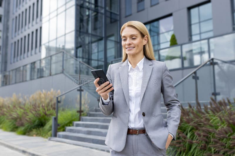 Confident businesswoman in a gray suit smiles as she reads a message on her smartphone, standing outside a modern office building. Confident businesswoman in a gray suit smiles as she reads a message on her smartphone, standing outside a modern office building.