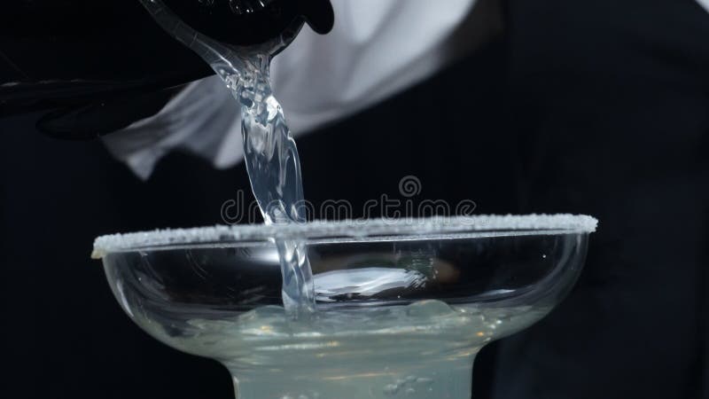 Professional bartender pouring fresh margarita with salted garnish glass. Macrography of skilled bartender hands prepare for making cocktail with black background at the bar. Slow motion. Comestible. Professional bartender pouring fresh margarita with salted garnish glass. Macrography of skilled bartender hands prepare for making cocktail with black background at the bar. Slow motion. Comestible.