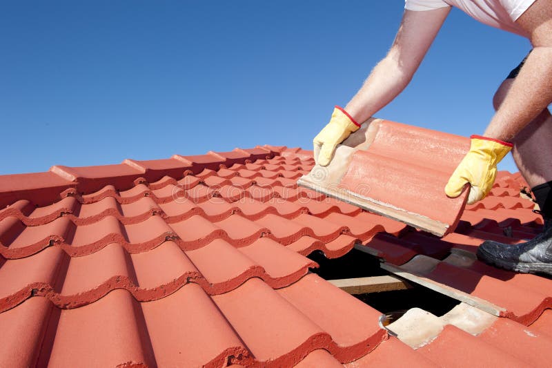 Roof repair, worker with yellow gloves replacing red tiles or shingles on house with blue sky as background and copy space. Roof repair, worker with yellow gloves replacing red tiles or shingles on house with blue sky as background and copy space.