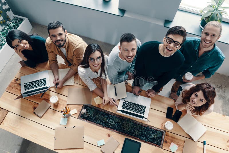 Successful professionals. Top view of young modern people in smart casual wear looking at camera and smiling while working in the creative office. Successful professionals. Top view of young modern people in smart casual wear looking at camera and smiling while working in the creative office