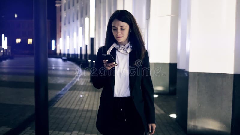 Επιχειρηματίας που περπατά σε μια πόλη νύχτας που παίρνει το smartphone και το χαμόγελό της