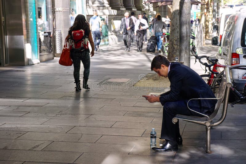 Επιχειρηματίες και χίπης στην οδό της Μελβούρνης. Οι νεολαίες έντυσαν καλά τη συνεδρίαση εργαζομένων γραφείων σε έναν πάγκο εξετάζοντας το τηλέφωνό του στην πόλη Αυστραλία της Μελβούρνης με έναν χίπη που περπατά από μπροστά
