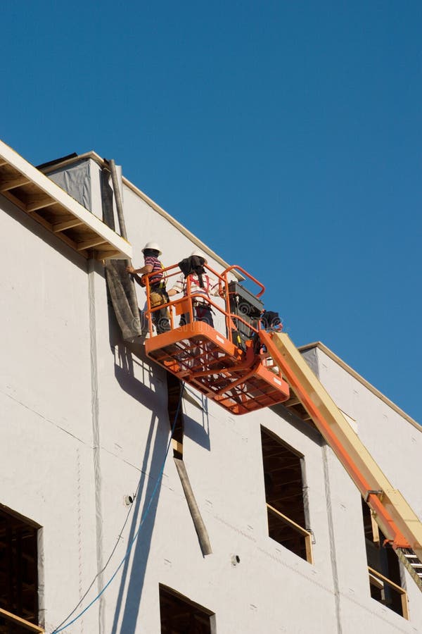 Two construction workers on a cherry picker working on a building under construction. Worker faces are hidden and not recognizable in the image. Two construction workers on a cherry picker working on a building under construction. Worker faces are hidden and not recognizable in the image.