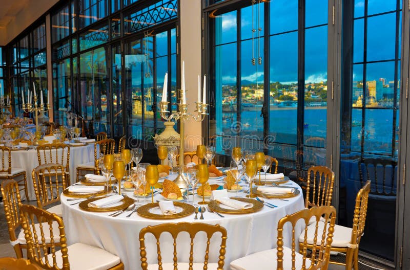 Wedding, party or business event tables decoration in white and golden tones. Silvery candelabrums and cutlery. Beautiful wide windows with urban bay view. Wedding, party or business event tables decoration in white and golden tones. Silvery candelabrums and cutlery. Beautiful wide windows with urban bay view.