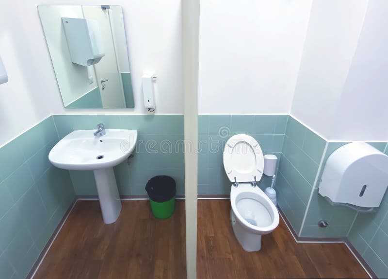 Wide angle view of a toilet with the wooden floor and tiled wall with a mirror, a green basket, a sink and the ceramic bowl. Wide angle view of a toilet with the wooden floor and tiled wall with a mirror, a green basket, a sink and the ceramic bowl