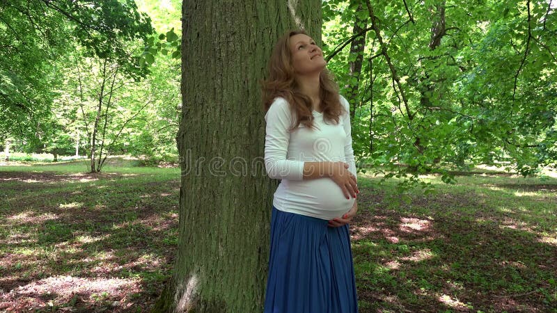 Ευτυχές καυκάσιο έγκυο κορίτσι που κτυπά την κοιλιά της που στέκεται κοντά στο μεγάλο κορμό δέντρων