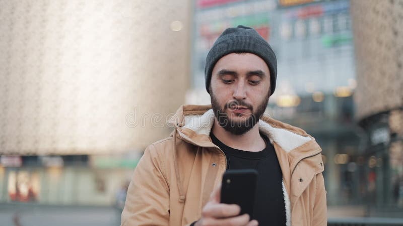Ευτυχές νέο άτομο γενειάδων που χρησιμοποιεί το smartphone που περπατά στη λεωφόρο πλησίον αγορών οδών Φορά ένα σακάκι φθινοπώρου