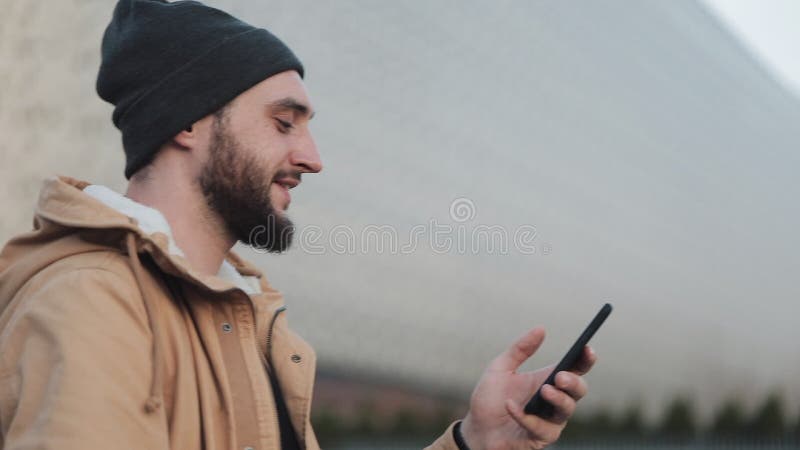 Ευτυχές νέο άτομο γενειάδων που χρησιμοποιεί το smartphone που περπατά στη λεωφόρο πλησίον αγορών οδών Φορά ένα σακάκι φθινοπώρου