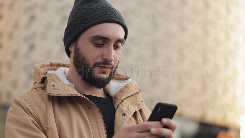 Ευτυχές νέο άτομο γενειάδων που χρησιμοποιεί το smartphone στη λεωφόρο πλησίον αγορών οδών Φορά ένα σακάκι φθινοπώρου και ένα πλε