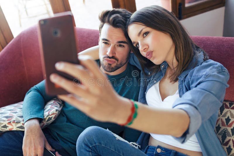 Ευτυχές νέο ζεύγος που παίρνει ένα selfie με το έξυπνο τηλέφωνο καθμένος στον καναπέ στο σπίτι στοκ εικόνα Ευτυχές νέο ζεύγος που παίρνει ένα selfie με το έξυπνο τηλέφωνο καθμένος στον καναπέ στο σπίτι στοκ εικόνα