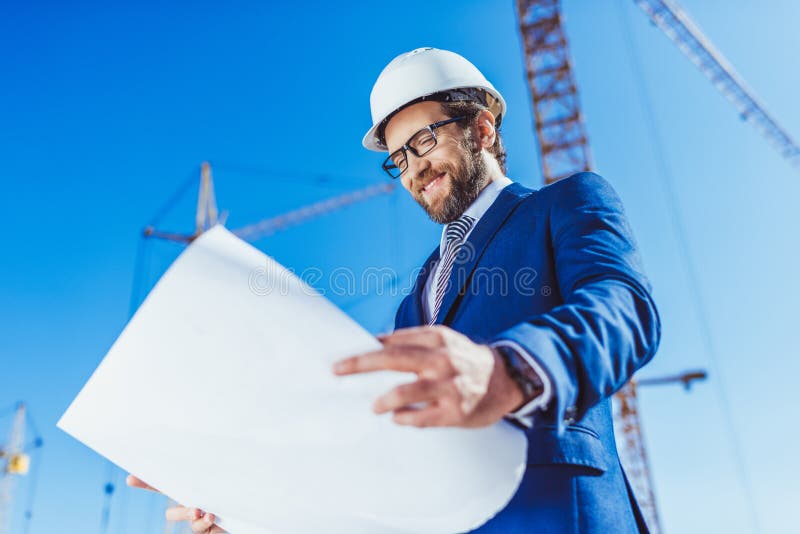 Ευτυχής επιχειρηματίας hardhat και κοστούμι που εξετάζει την οικοδόμηση των σχεδίων στοκ φωτογραφία Ευτυχής επιχειρηματίας hardhat και κοστούμι που εξετάζει την οικοδόμηση των σχεδίων στοκ φωτογραφία