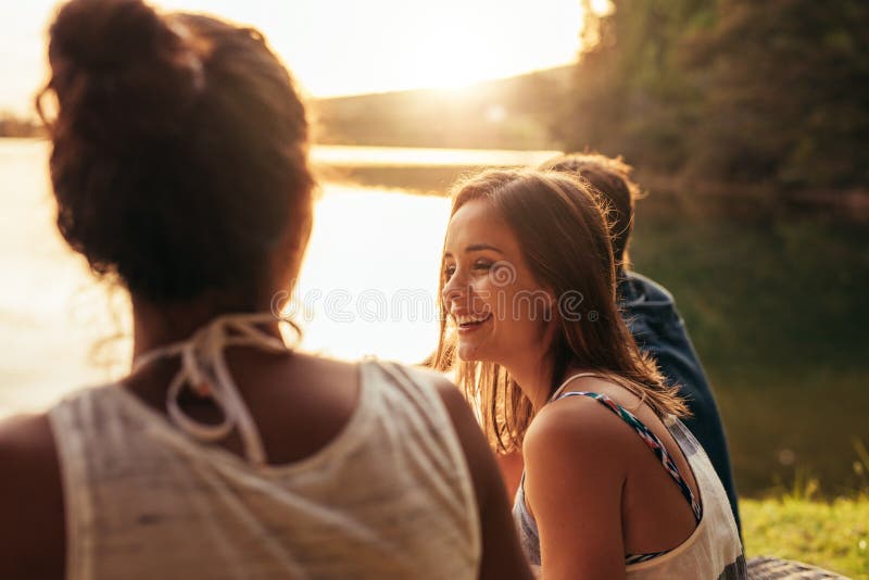 Ευτυχής νέα συνεδρίαση γυναικών από μια λίμνη με τους φίλους της στοκ φωτογραφία Ευτυχής νέα συνεδρίαση γυναικών από μια λίμνη με τους φίλους της στοκ φωτογραφία
