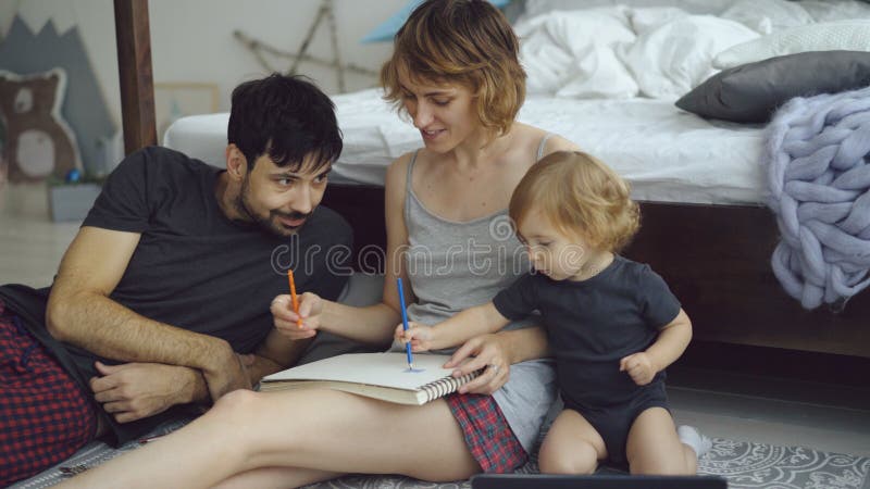 Ευτυχής οικογένεια με το χαριτωμένο λατρευτό σχέδιο κορών στο λεύκωμα με τα μολύβια που κάθεται κοντά στο κρεβάτι στο σπίτι στοκ φωτογραφίες με δικαίωμα ελεύθερης χρήσης Ευτυχής οικογένεια με το χαριτωμένο λατρευτό σχέδιο κορών στο λεύκωμα με τα μολύβια που κάθεται κοντά στο κρεβάτι στο σπίτι στοκ φωτογραφίες με δικαίωμα ελεύθερης χρήσης