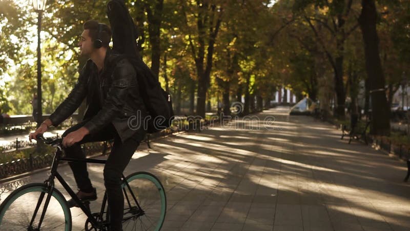 Ευτυχής, χαμογελώντας νεαρός άνδρας στα μαύρα περιστασιακά ενδύματα, ακουστικά που οδηγούν ένα ποδήλατο από το στρωμένο πάρκο πόλ