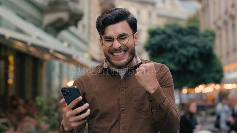 Happy hopeful lucky Indian Arabian ethnic man guy businessman male young student hold mobile phone smartphone wishing hope praying crossed fingers winner yes scream victory gesture outside city street. High quality 4k footage. Happy hopeful lucky Indian Arabian ethnic man guy businessman male young student hold mobile phone smartphone wishing hope praying crossed fingers winner yes scream victory gesture outside city street. High quality 4k footage