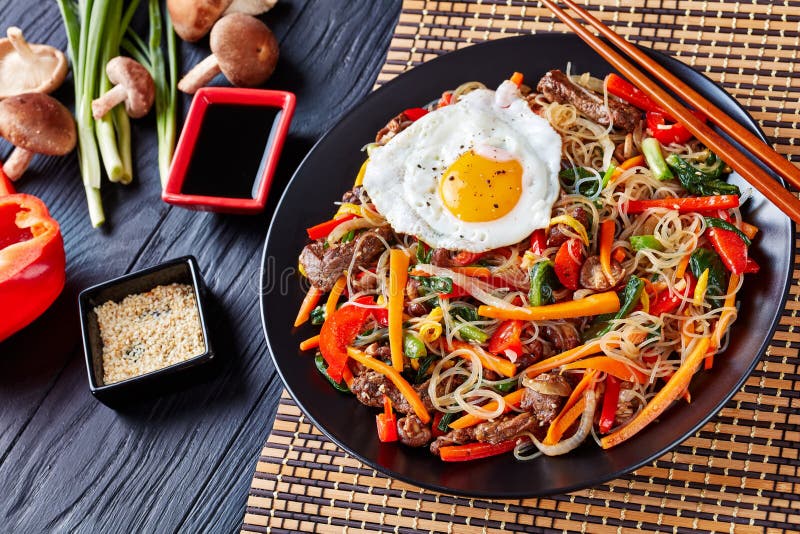 Delicious korean dish Japchae: mix of glass noodles, beef and stir fried onion, bell pepper, garlic, spinach, carrot, shiitake mushrooms with sesame seeds oil and soy sauce, on a black plate. Delicious korean dish Japchae: mix of glass noodles, beef and stir fried onion, bell pepper, garlic, spinach, carrot, shiitake mushrooms with sesame seeds oil and soy sauce, on a black plate