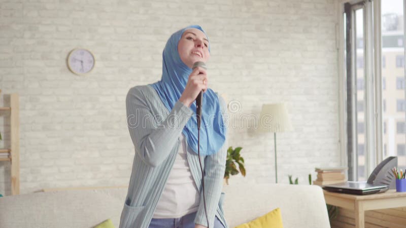 Εύθυμο εκφραστικό μουσουλμανικό κορίτσι στο hijab στο μικρόφωνο καραόκε στο σπίτι