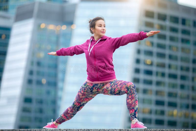 Young attractive woman practicing yoga, standing in Warrior two exercise, Virabhadrasana II pose, working out wearing pink sportswear, outdoor full length, modern skyscraper background. Young attractive woman practicing yoga, standing in Warrior two exercise, Virabhadrasana II pose, working out wearing pink sportswear, outdoor full length, modern skyscraper background