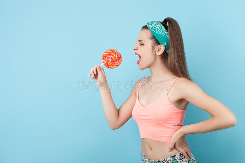 Beautiful hipster girl is standing and holding a lollipop. She is shouting at it as if it was a microphone. The girl is looking at sweet food with aggression. Isolated and copy space in left side. Beautiful hipster girl is standing and holding a lollipop. She is shouting at it as if it was a microphone. The girl is looking at sweet food with aggression. Isolated and copy space in left side