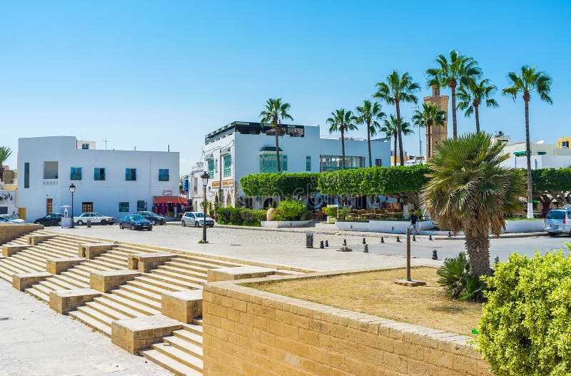 MAHDIA, TUNISIA - AUGUST 29, 2015: The scenic cityscape with lush greenery of ornamental park, quarters of old town with white housing, tourist cafes in shade of trees and minaret of Soliman Hamza mosque on background, on August 29 in Mahdia. MAHDIA, TUNISIA - AUGUST 29, 2015: The scenic cityscape with lush greenery of ornamental park, quarters of old town with white housing, tourist cafes in shade of trees and minaret of Soliman Hamza mosque on background, on August 29 in Mahdia.