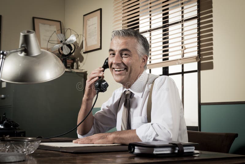 Smiling handsome businessman on the phone working at desk, 1950s vintage office. Smiling handsome businessman on the phone working at desk, 1950s vintage office.