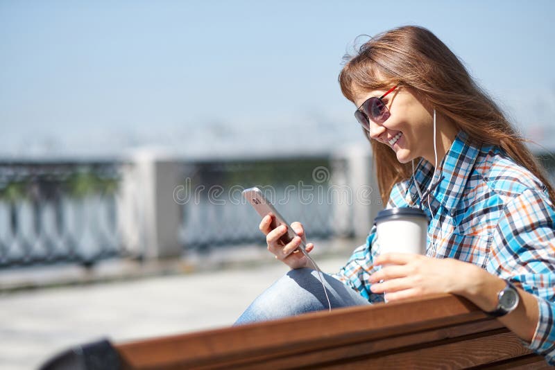 Η νέα ευτυχής γυναίκα χρησιμοποιεί το smartphone Κρατά ένα φλιτζάνι του καφέ εγγράφου στοκ φωτογραφίες με δικαίωμα ελεύθερης χρήσης Η νέα ευτυχής γυναίκα χρησιμοποιεί το smartphone Κρατά ένα φλιτζάνι του καφέ εγγράφου στοκ φωτογραφίες με δικαίωμα ελεύθερης χρήσης