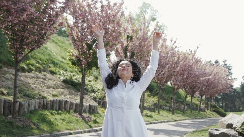 Η νέα ξένοιαστη γυναίκα που φορά το άσπρο μακρύ φόρεμα απολαμβάνει των ανθίζοντας δέντρων sakura