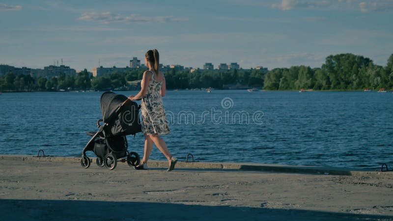 Η νέα όμορφη μητέρα στο φόρεμα περπατά κοντά στον ποταμό με τον περιπατητή