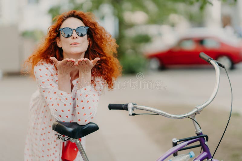 Η στοργική redhead γυναίκα στέλνει το φιλί αέρα στον εραστή στην οδό, κρατά τους φοίνικες κοντά στο στόμα, φορά γύρω από τις σκιέ στοκ φωτογραφία Η στοργική redhead γυναίκα στέλνει το φιλί αέρα στον εραστή στην οδό, κρατά τους φοίνικες κοντά στο στόμα, φορά γύρω από τις σκιέ στοκ φωτογραφία