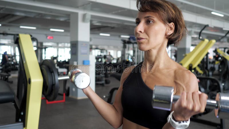 Η φίλαθλη ώριμη γυναίκα κάνει την άσκηση δικέφαλων μυών με τους αλτήρες στη γυμναστική