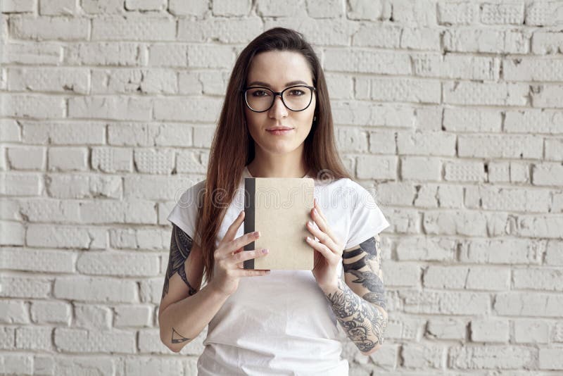 Η όμορφη διαστισμένη hipster γυναίκα θέτει στην άσπρη μπλούζα, που απομονώνεται στον άσπρο τουβλότοιχο, κρατά ένα βιβλίο διαθέσιμ στοκ εικόνες με δικαίωμα ελεύθερης χρήσης Η όμορφη διαστισμένη hipster γυναίκα θέτει στην άσπρη μπλούζα, που απομονώνεται στον άσπρο τουβλότοιχο, κρατά ένα βιβλίο διαθέσιμ στοκ εικόνες με δικαίωμα ελεύθερης χρήσης