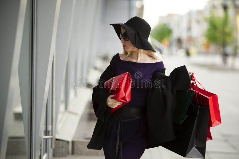 Η όμορφη νέα μοντέρνη ξανθή γυναίκα στο μπλε φόρεμα, το παλτό, τα γυαλιά ηλίου και το καπέλο με τις αγορές τοποθετεί το περπάτημα στοκ εικόνα Η όμορφη νέα μοντέρνη ξανθή γυναίκα στο μπλε φόρεμα, το παλτό, τα γυαλιά ηλίου και το καπέλο με τις αγορές τοποθετεί το περπάτημα στοκ εικόνα