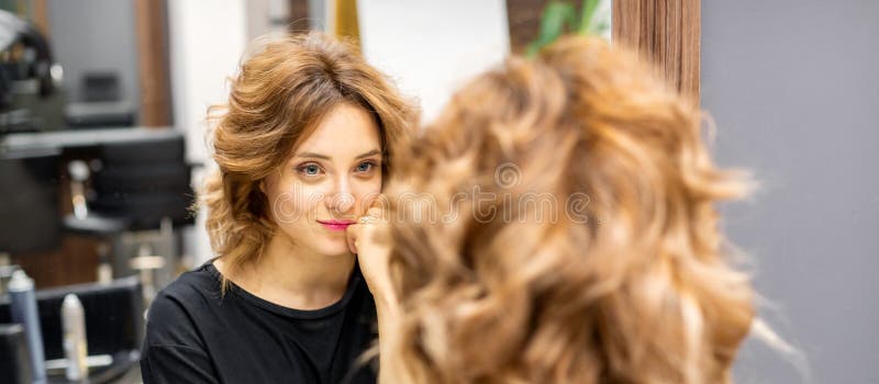η όμορφη νεαρή καυκάσια κοκκινομάλλα γυναίκα με ένα νέο κοντό κυματιστό στυλ που κοιτά στον καθρέφτη κοιτάζοντας το στυλ του στοκ εικόνα