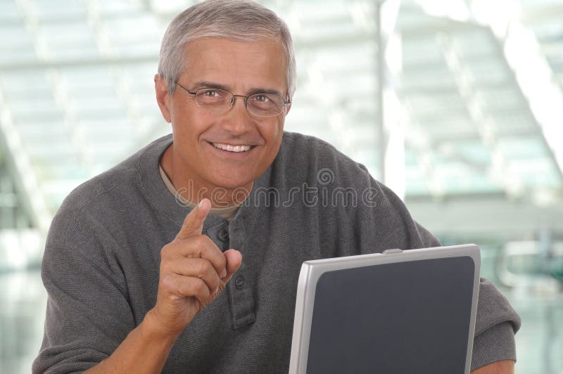 Middle aged man sitting at laptop computer and pointing at camera. Man is smiling and casually dressed. Horizontal format with blurred office background. Middle aged man sitting at laptop computer and pointing at camera. Man is smiling and casually dressed. Horizontal format with blurred office background.