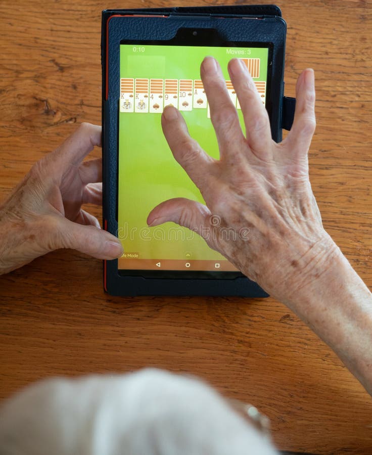 An elderly gray-haired woman playing solitaire on a tablet. Her hands and part of her head are shown. Photographed from above. An elderly gray-haired woman playing solitaire on a tablet. Her hands and part of her head are shown. Photographed from above.