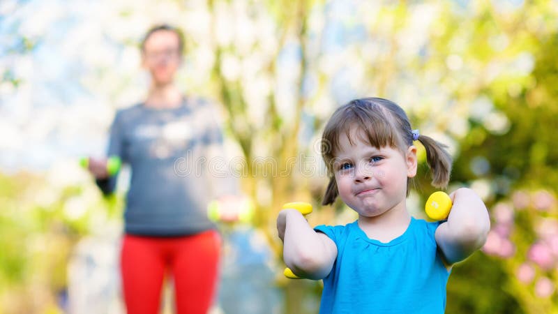 Happy little girl lifting dumbbells in front of her mother in park outdoors. Shallow depth of field. Creamy background. Happy little girl lifting dumbbells in front of her mother in park outdoors. Shallow depth of field. Creamy background.