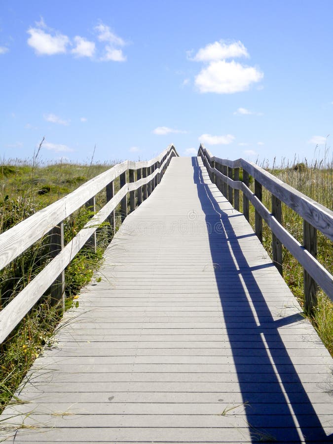 A vertical view of a long wooden boardwalk ocean beach access leading over sand dunes on a beautiful summer day in the Outer Banks of North Carolina. A vertical view of a long wooden boardwalk ocean beach access leading over sand dunes on a beautiful summer day in the Outer Banks of North Carolina.