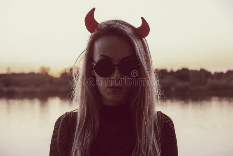 Deadlly serious female wearing sunglasses and Devils horns posing in front of sunset lake. Deadlly serious female wearing sunglasses and Devils horns posing in front of sunset lake
