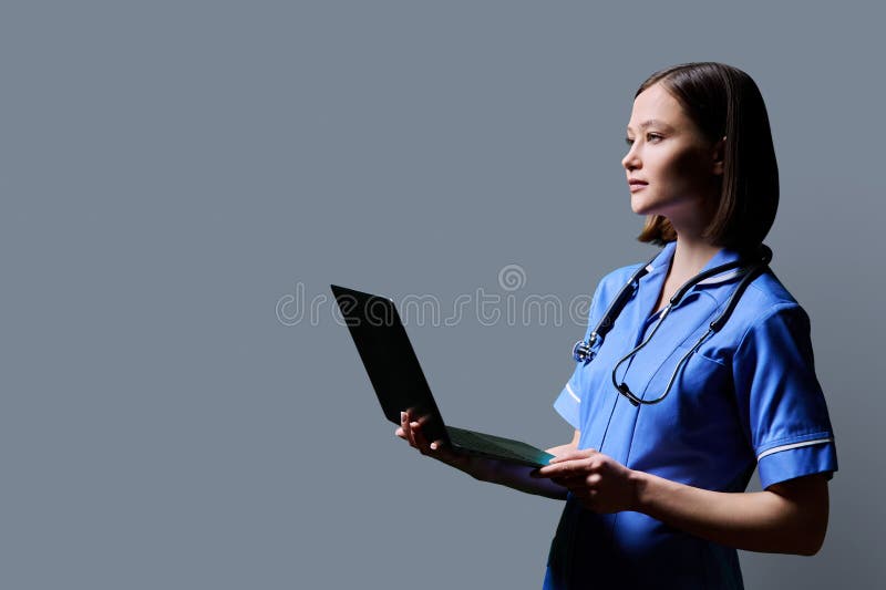 Serious young female nurse using laptop computer, profile view on gray studio background. Mobile apps applications technologies in medical services health professional assistance medical care concept. Serious young female nurse using laptop computer, profile view on gray studio background. Mobile apps applications technologies in medical services health professional assistance medical care concept