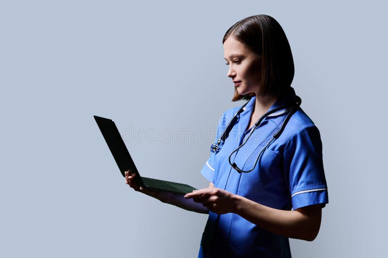 Serious young female nurse using laptop computer, profile view on gray studio background. Mobile apps applications technologies in medical services health professional assistance medical care concept. Serious young female nurse using laptop computer, profile view on gray studio background. Mobile apps applications technologies in medical services health professional assistance medical care concept