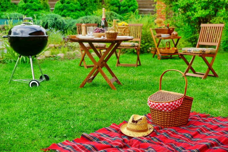 Closeup Of Red Picnic Blanket With Straw Hat And Basket Or Hamper. Blurred Outdoor Wooden Furniture In The Background. Family Home Backyard BBQ Party Or Picnic Conceptual Scene. Closeup Of Red Picnic Blanket With Straw Hat And Basket Or Hamper. Blurred Outdoor Wooden Furniture In The Background. Family Home Backyard BBQ Party Or Picnic Conceptual Scene