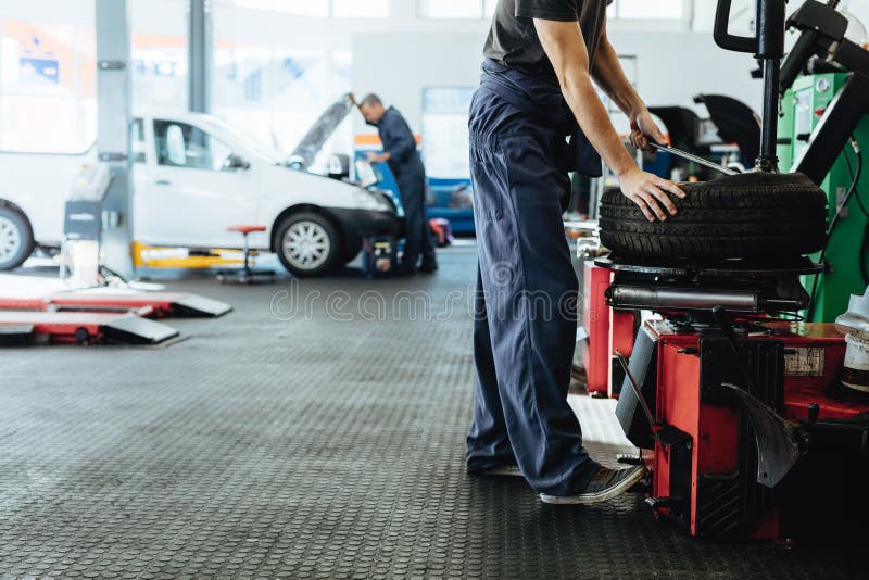 Car repair shop with mechanics working. Mechanic replacing tire of while on machine and other inspecting a vehicle. Car repair shop with mechanics working. Mechanic replacing tire of while on machine and other inspecting a vehicle.