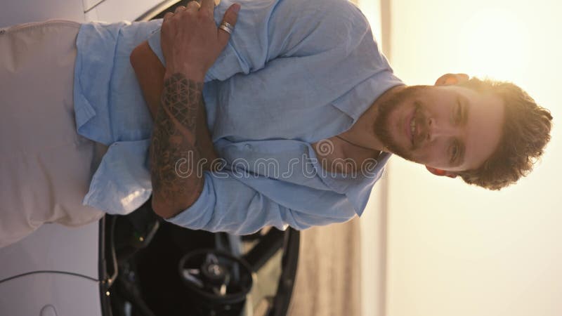 κατακόρυφο βίντεο : πορτρέτο ενός γενειοφόρου άντρα με μπλε πουκάμισο στέκεται με σταυρωμένα τα χέρια και ακουμπά σε ένα λευκό μετ