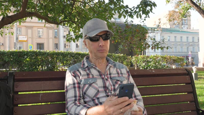 Καυκάσιος με γυαλιά ηλίου κάθεται στο πάρκο σε πάγκο τρώει κλικ στο smartphone απόθεμα βίντεο