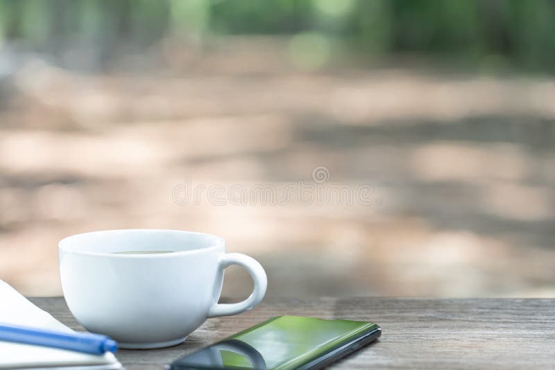 καυτή κούπα καφέ με λευκή χάρτινη πένα και κινητό τηλέφωνο σε ξύλινο τραπέζι και θέα στη φύση. καυτή κούπα καφέ με λευκή χάρτα και κινητό τηλέφωνο στο ξύλινο τραπέζι και θέα στη φύση στο αγροτικό χωριό