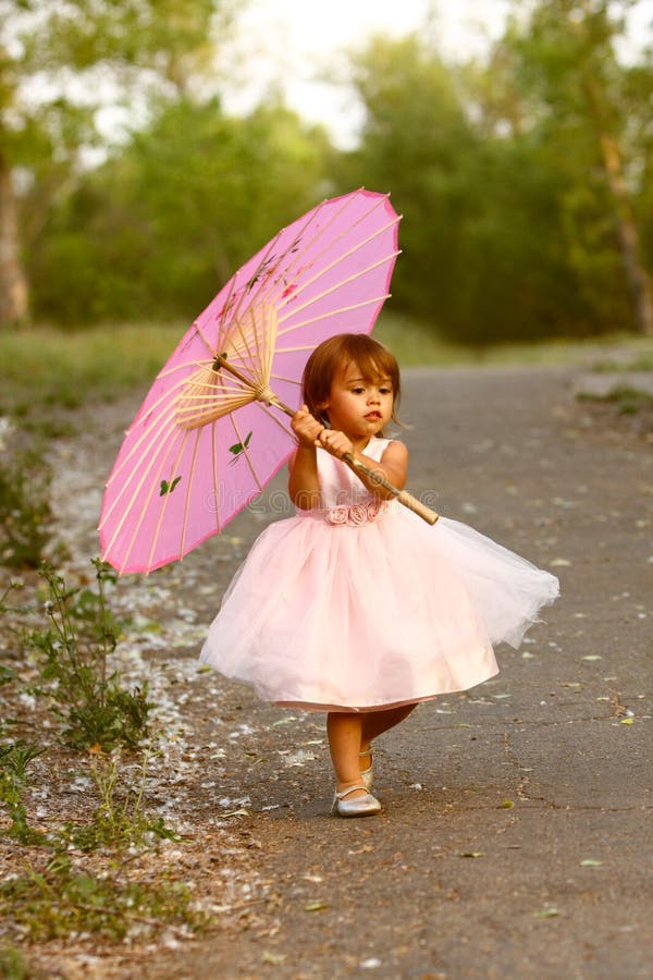 Cute, multi-racial, 2 year old girl walks in a park and carries a pink parasol. Little girl is Asian/Caucasian with brown hair and eyes. Girl is wearing a dressy, pale pink, full dress. Cute, multi-racial, 2 year old girl walks in a park and carries a pink parasol. Little girl is Asian/Caucasian with brown hair and eyes. Girl is wearing a dressy, pale pink, full dress.