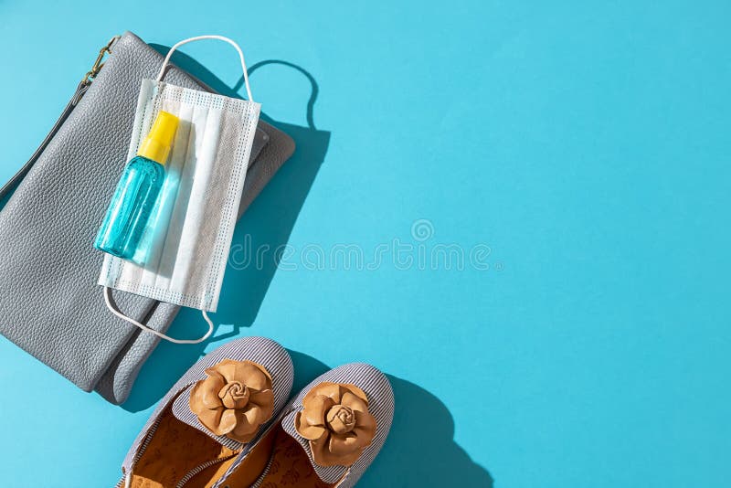 Stylish female shoes and Leather bag out with face mask ,sanitizer hand gel to protect from Coronavirus or COVID-19 on a blue background with sunlight shadow. Copy space, flat lay. Stylish female shoes and Leather bag out with face mask ,sanitizer hand gel to protect from Coronavirus or COVID-19 on a blue background with sunlight shadow. Copy space, flat lay