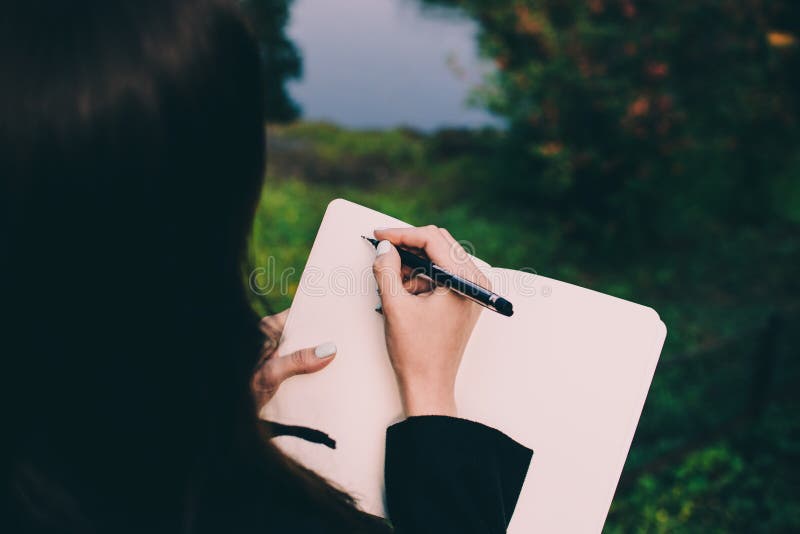 Girl writing down her thoughts in a notebook against tiny blurred swan house in a middle of a pond in a park. Pale hand and colorful background. Girl writing down her thoughts in a notebook against tiny blurred swan house in a middle of a pond in a park. Pale hand and colorful background