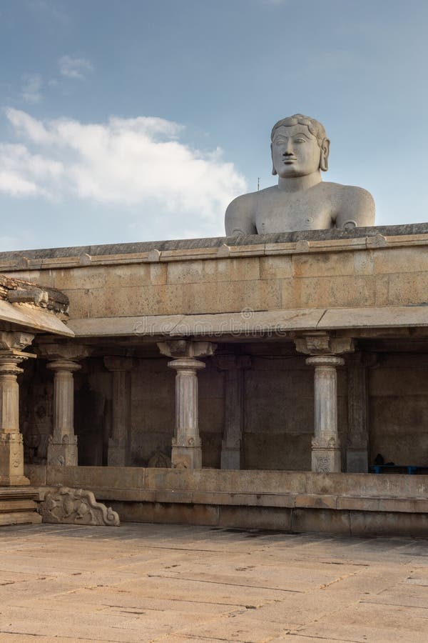 Shravanabelagola, Karnataka, India - November 1, 2013: At the Jain Tirth, gray granite Giant Bhagwan Bahubali puts torso above brown stone temple building against light blue sky. Shravanabelagola, Karnataka, India - November 1, 2013: At the Jain Tirth, gray granite Giant Bhagwan Bahubali puts torso above brown stone temple building against light blue sky.