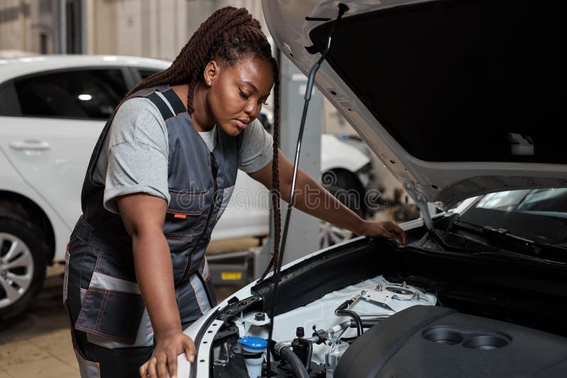 Tired african auto mechanic woman looking to understand what is wrong with car, looking inside of hood, side view portrait. Young female in auto service. copy space. Tired african auto mechanic woman looking to understand what is wrong with car, looking inside of hood, side view portrait. Young female in auto service. copy space